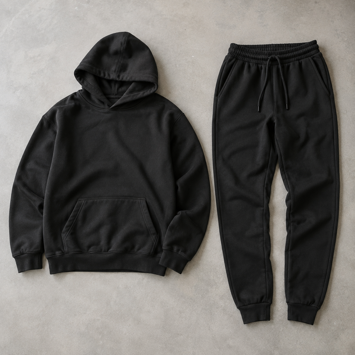 a coordinated black men's two piece outfit consisting of a plain long sleeve pullover hoodie with kangaroo pocket and matching full length cuffed drawstring joggers, both fully visible arranged side by side